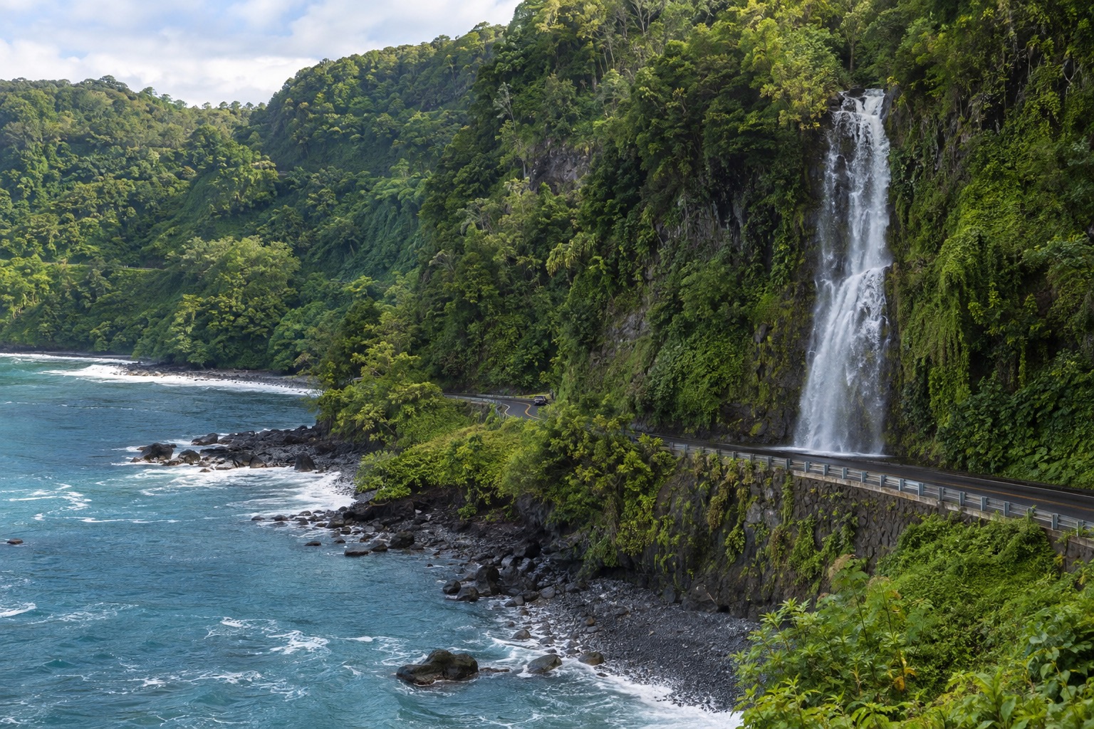 A waterfall dropping through dense green cliffs beside a narrow coastal highway