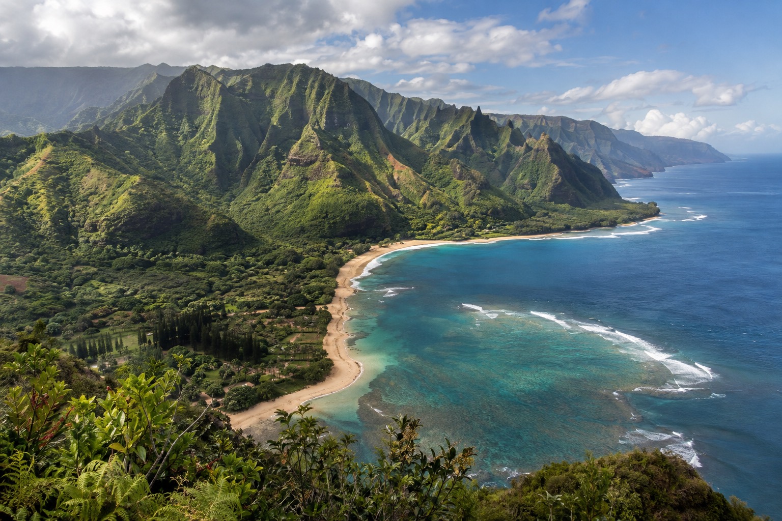 Green volcanic ridges above a curving sandy bay on Kauai under scattered clouds