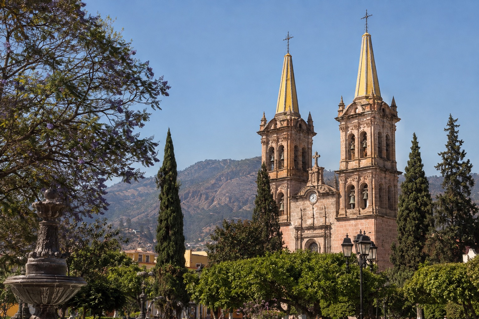 Twin yellow-tiled cathedral spires rising above a plaza with trees under a clear sky