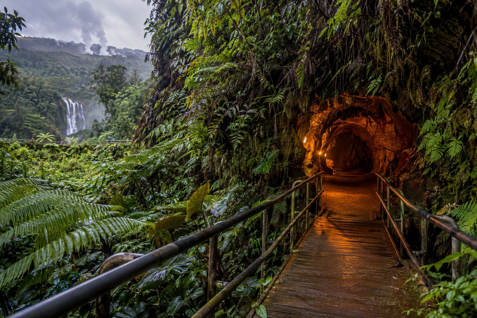 A boardwalk trail leading into the lit tunnel entrance of a lava tube surrounded by ferns
