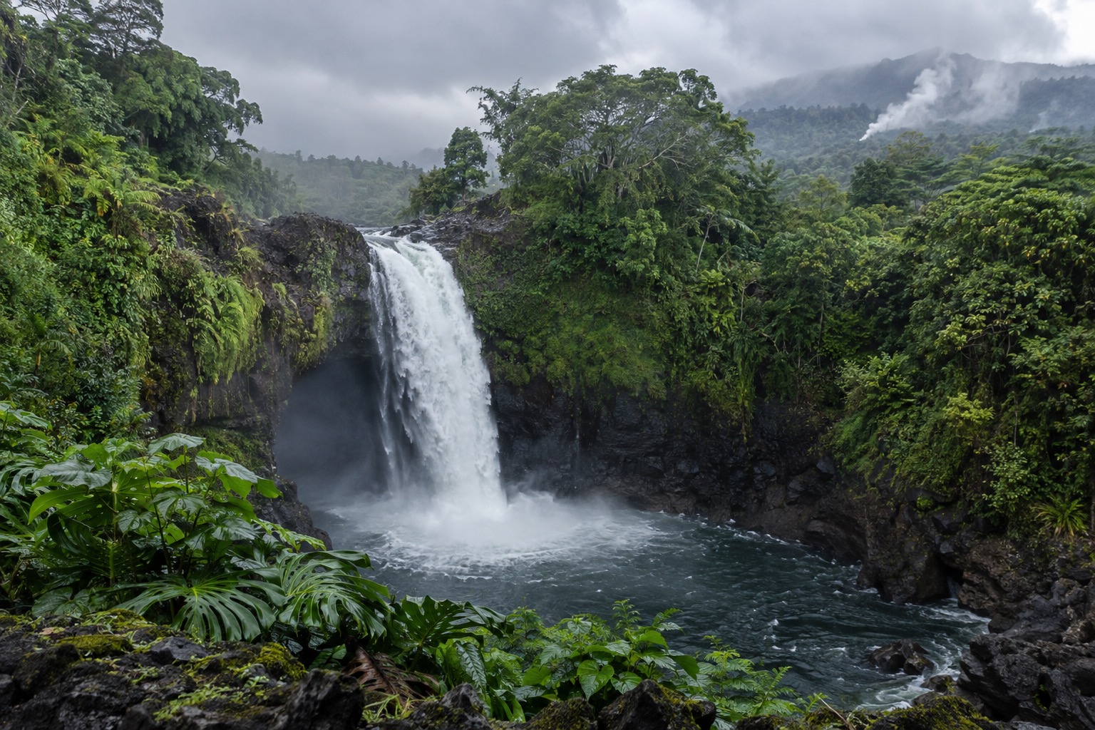 Misty rainforest waterfall falling into a rocky pool near Hilo, Hawaii, under an overcast sky