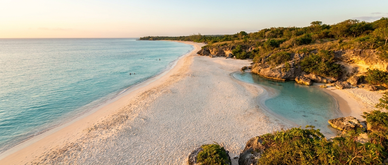 A long white-sand beach curving into calm turquoise water on one side and a rocky green cove on the other, golden afternoon light