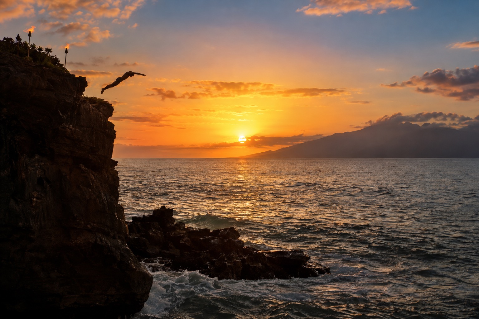 A torch-lit cliff at the north end of a Maui beach with a diver silhouetted against the sunset