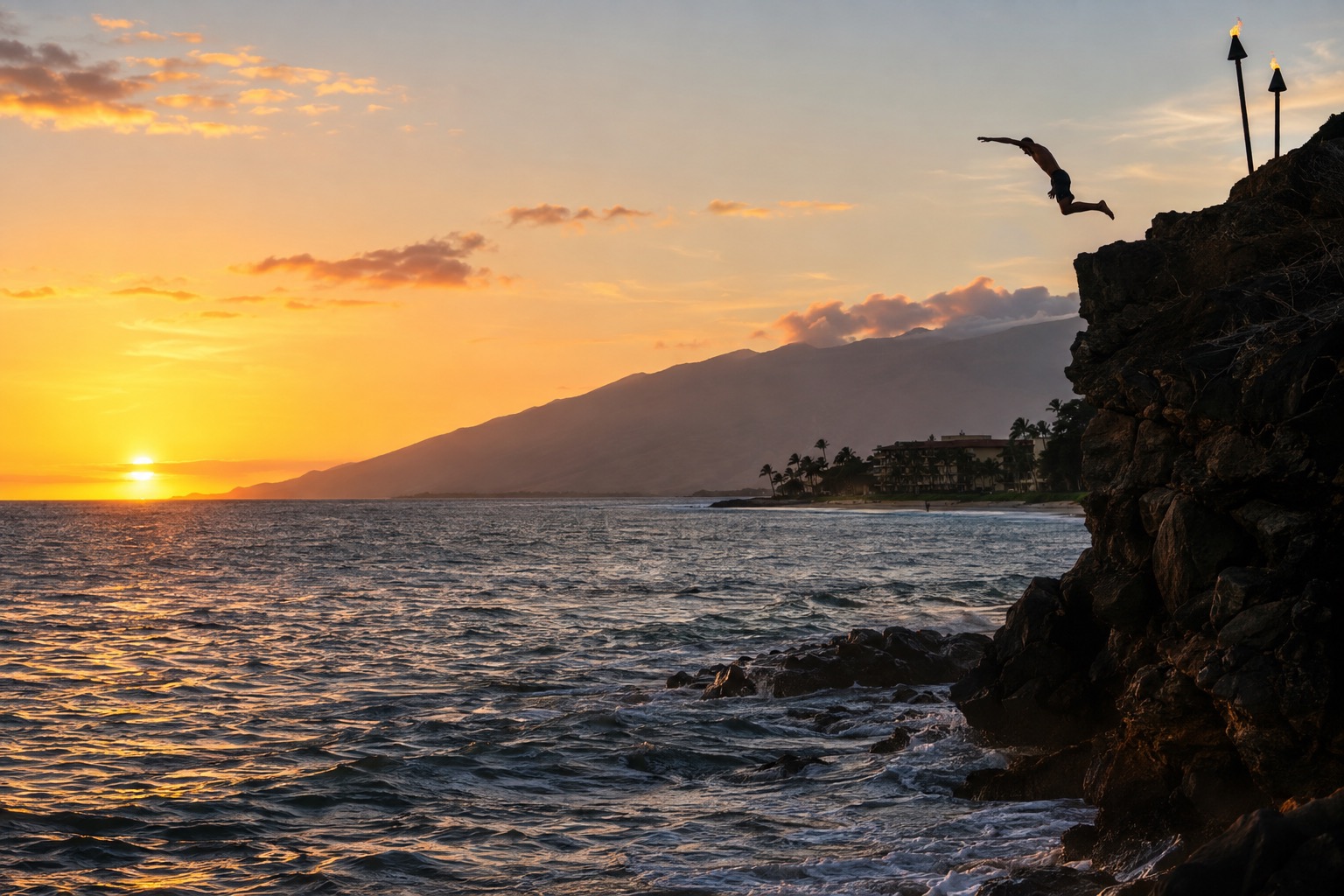 A torch-lit cliff at the north end of a beach at sunset with a diver mid-leap