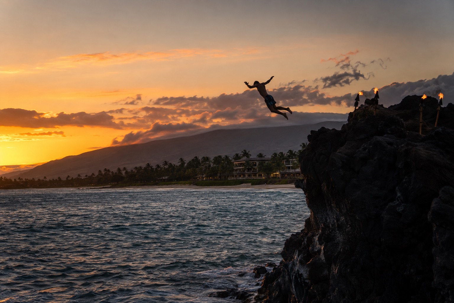 A cliff diver mid-leap off a dark lava outcrop into the ocean at dusk, torches lit along the rock