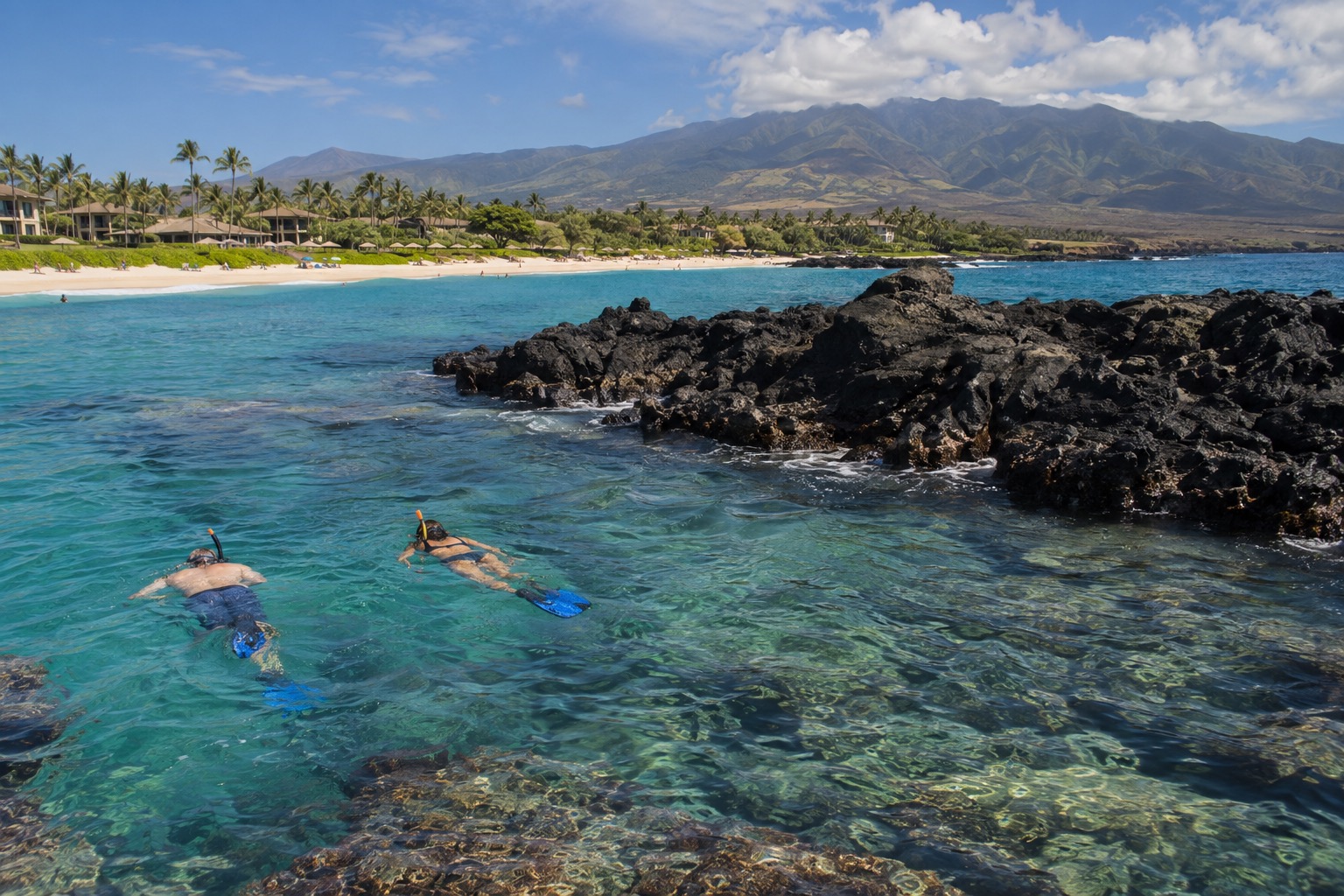 Snorkelers in clear water near a black lava outcrop off a white sand beach