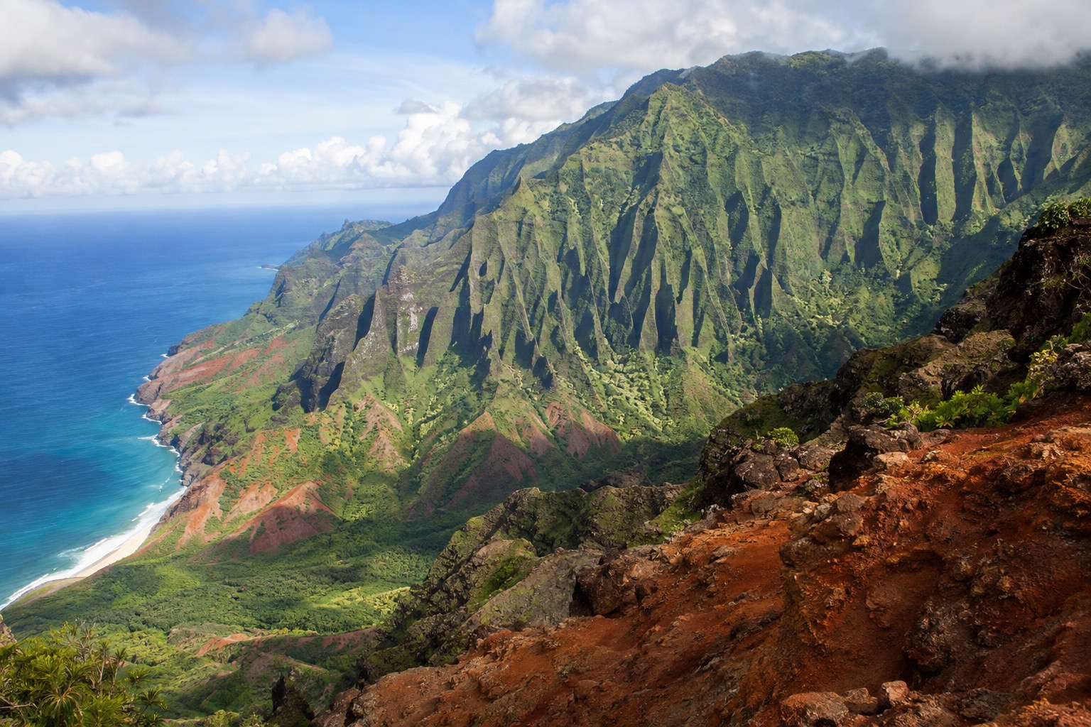 Steep red and green canyon walls under a partly cloudy Kauai sky