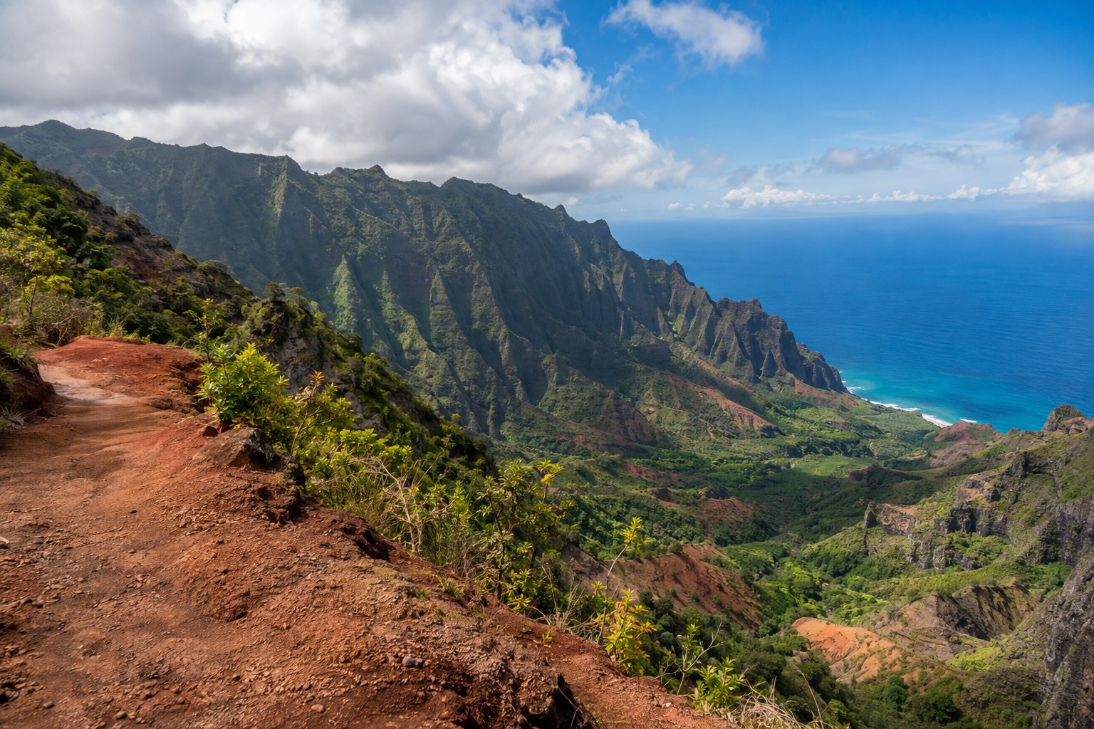 A dirt overlook road above Waimea Canyon's red cliffs under a partly cloudy Kauai sky