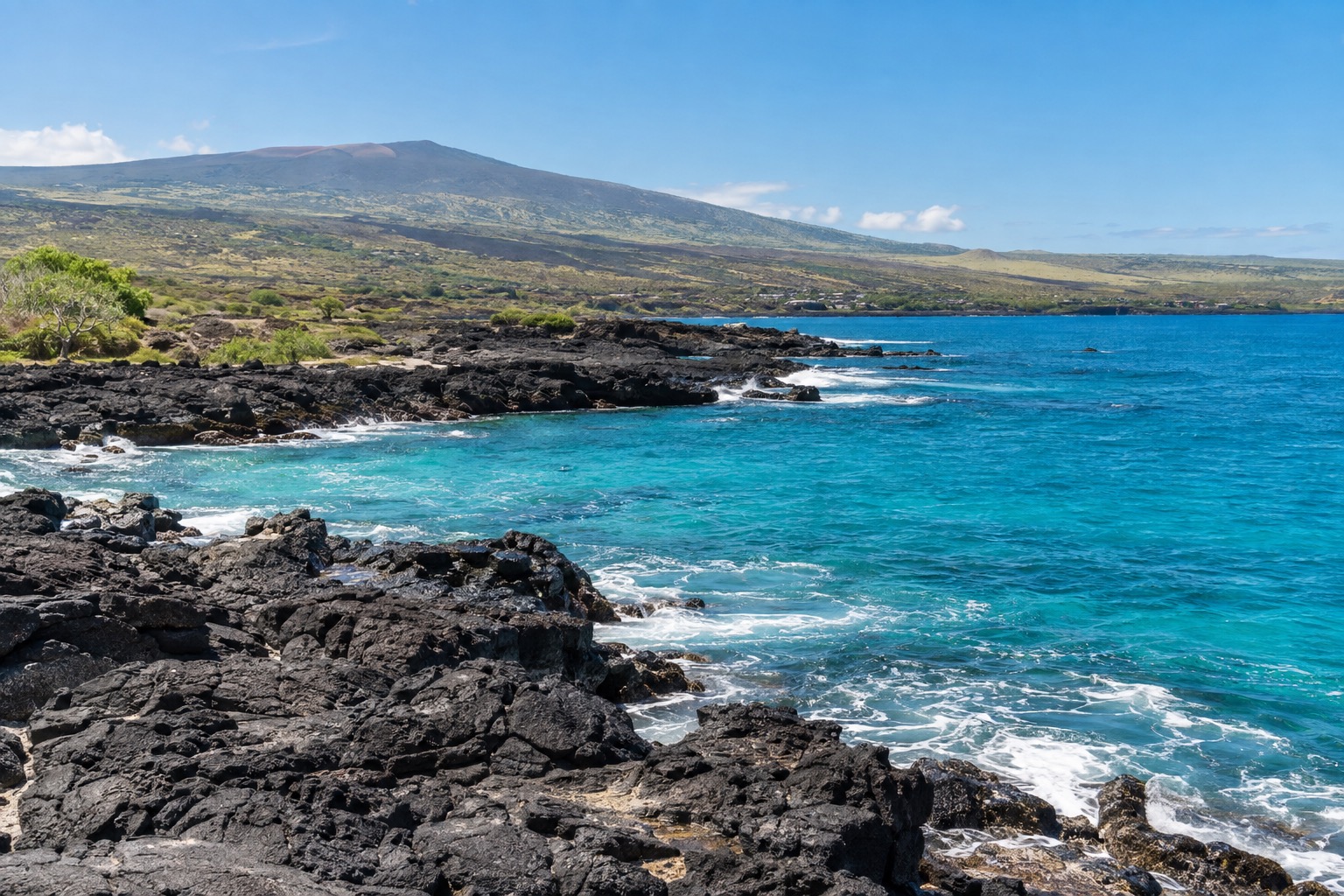 A rocky lava-rock shoreline in Kona, Hawaii, with turquoise water and a distant cinder cone hill under a clear sky