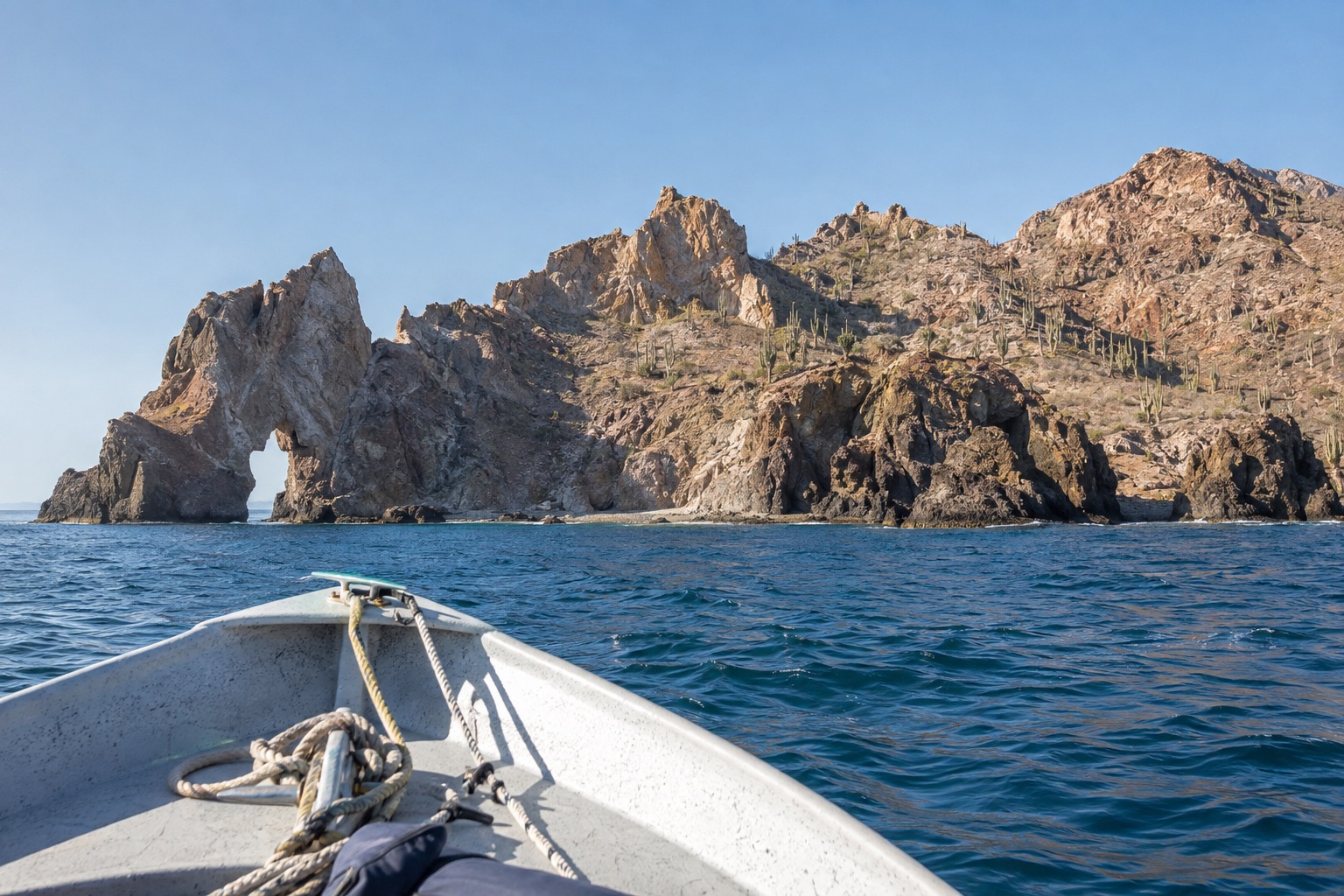 A small open boat crossing blue water toward a rugged desert island under a clear sky