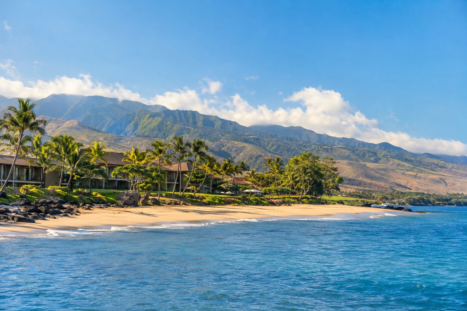 A crescent of white sand beach lined with palm trees and a low resort building on Maui's leeward coast, calm blue water in the foreground