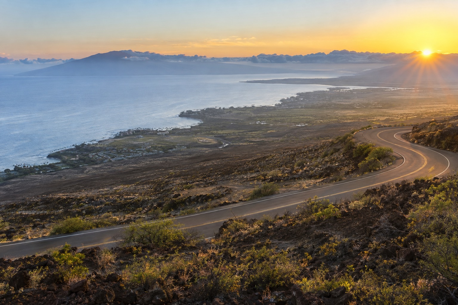 A paved road switchbacking up a volcanic slope above a layer of clouds at sunrise
