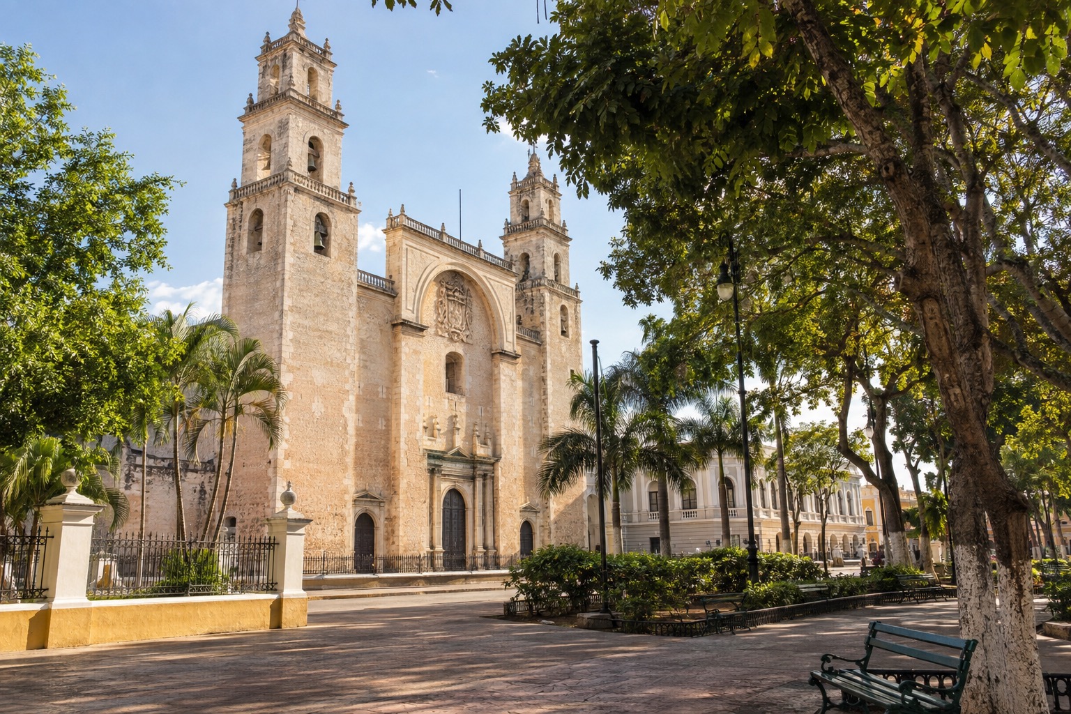 Sunlit stone facade of a colonial cathedral beside a tree-shaded plaza in Merida, Yucatan