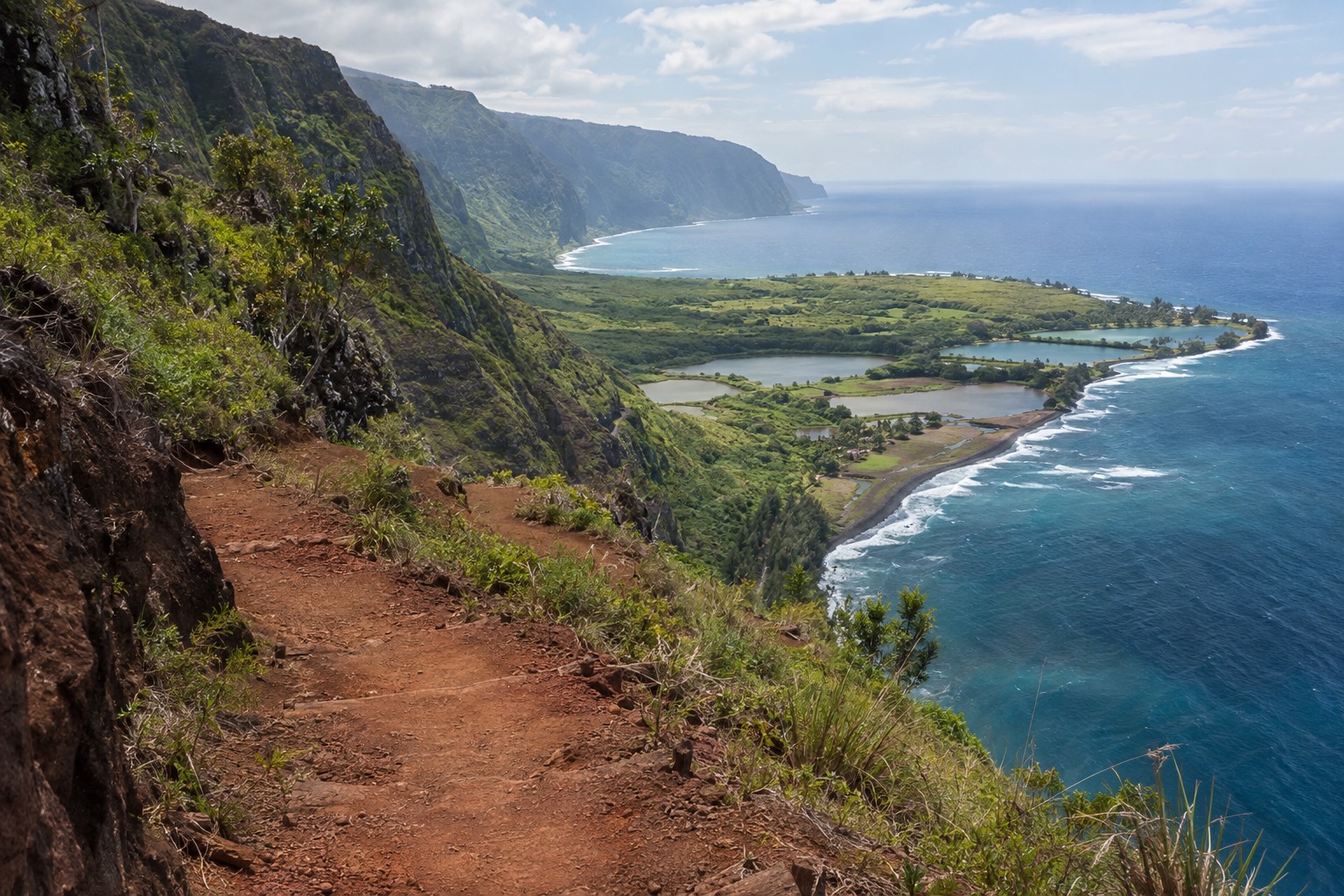 A red-dirt trail switchbacking down steep sea cliffs toward a green peninsula on Molokai's north shore