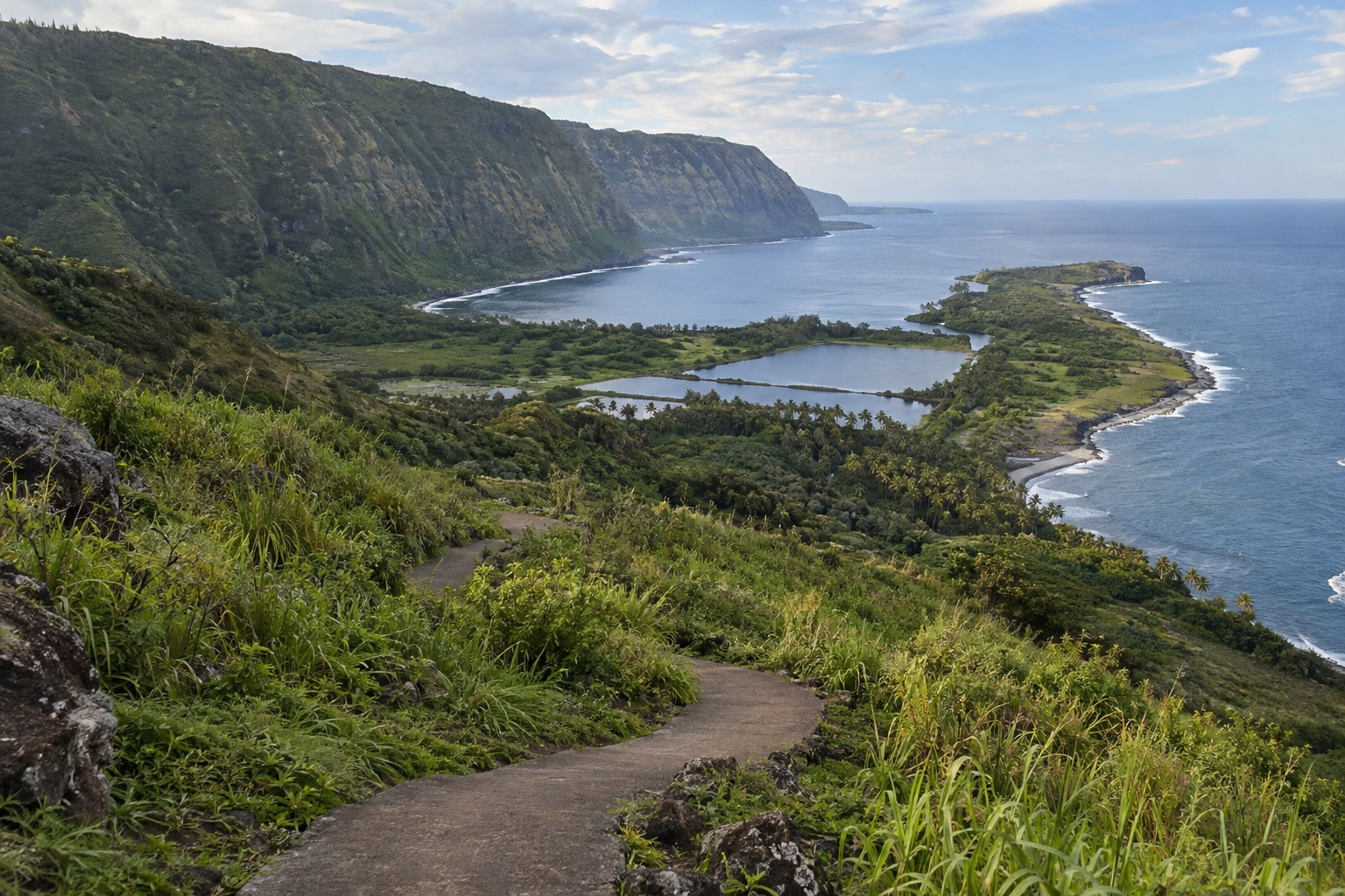 A paved trail switchbacking down a green sea cliff toward a narrow peninsula on Molokai