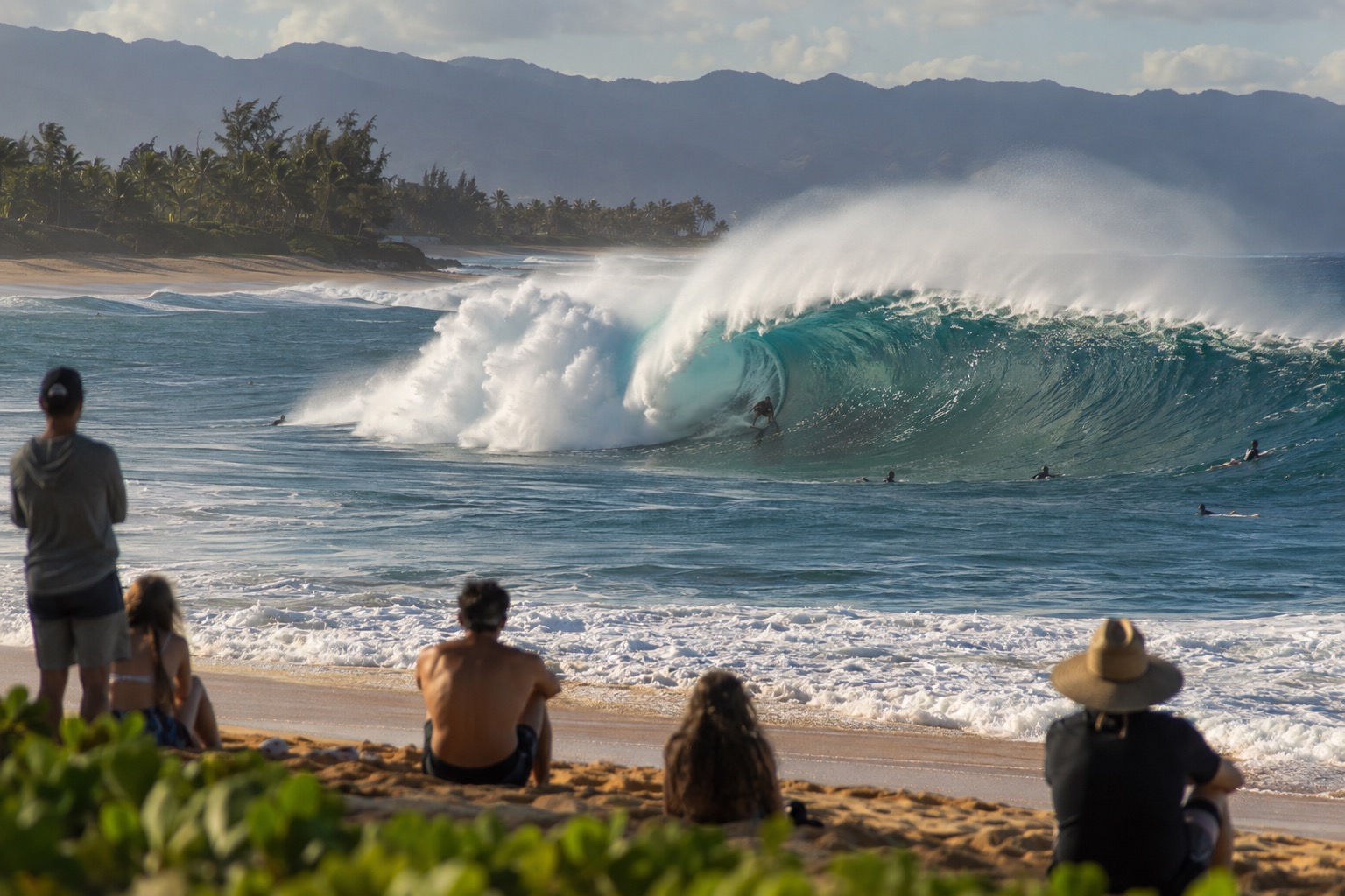 A large winter wave breaking over a shallow reef on Oahu's North Shore with beachgoers watching from the sand