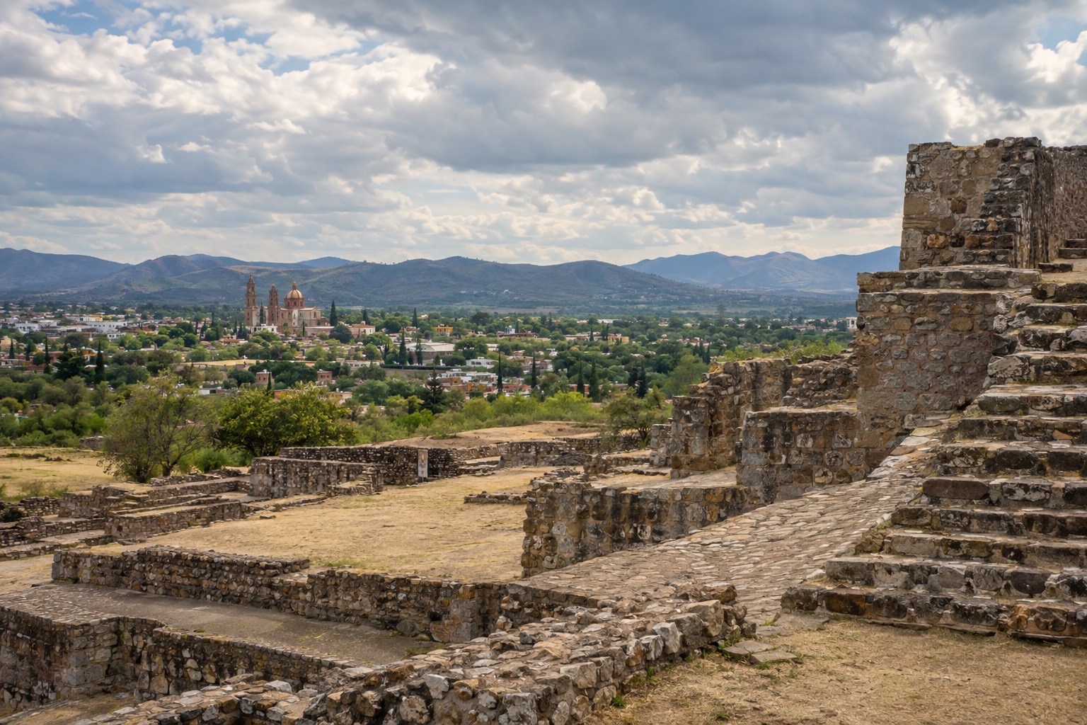 Stone platforms and stairways of an ancient hilltop ruin above a green valley under a cloudy sky