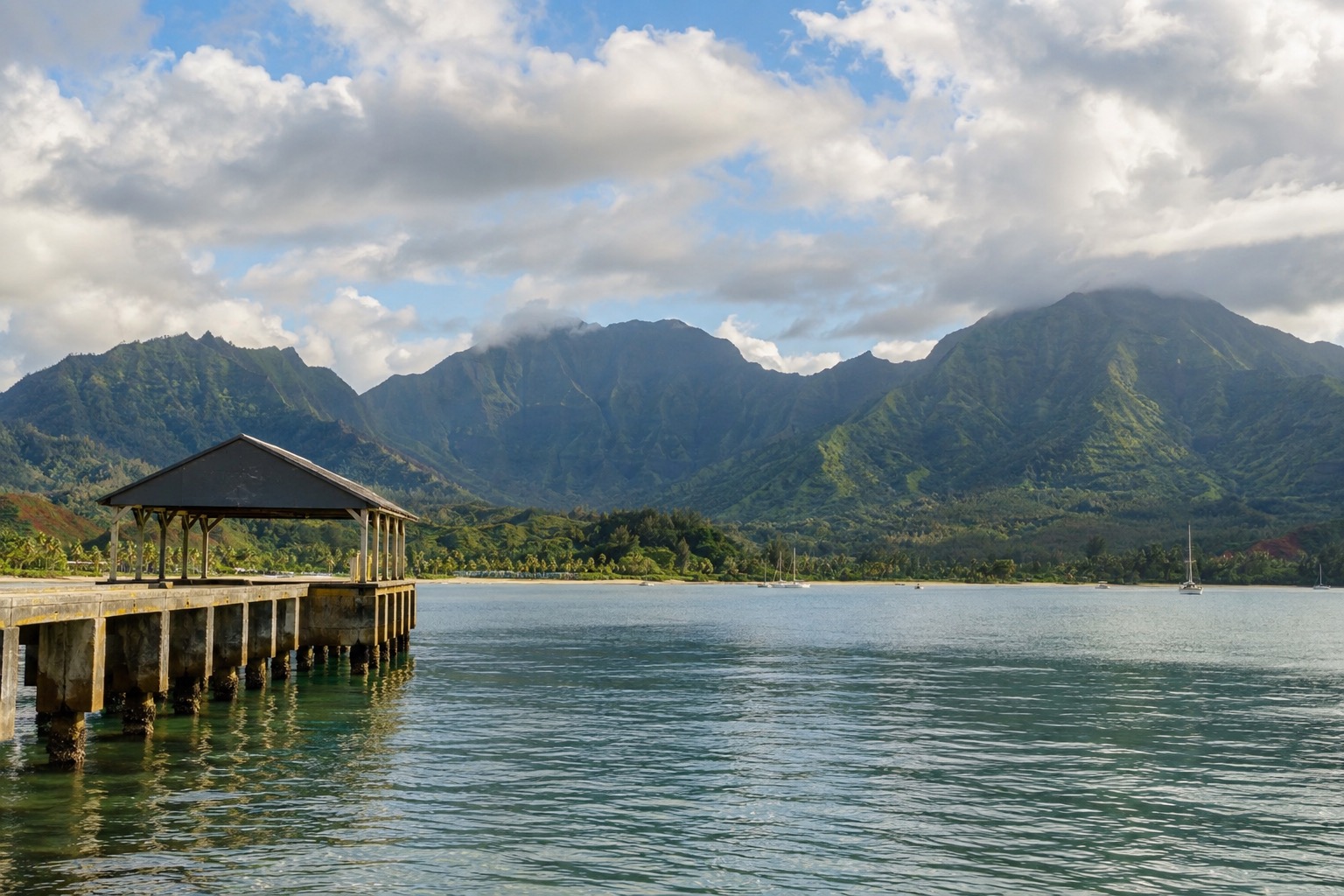 Calm water in Hanalei Bay seen from the pier, with green mountains under a partly cloudy sky