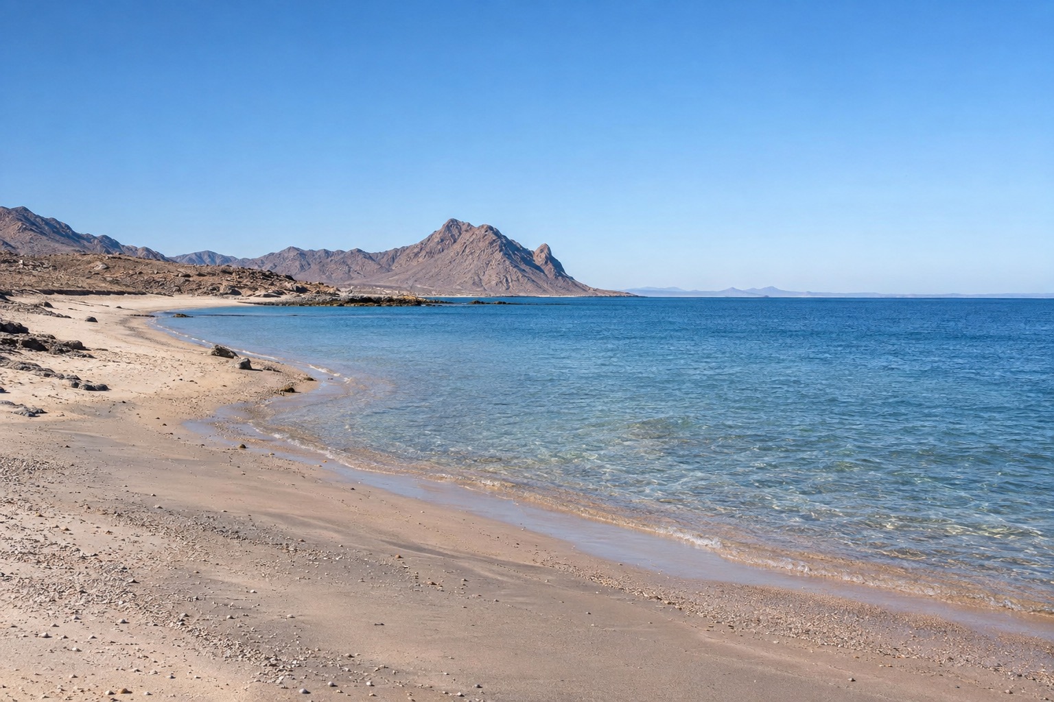 Wide sandy beach meeting calm blue water under a clear sky, with a low rocky point in the distance