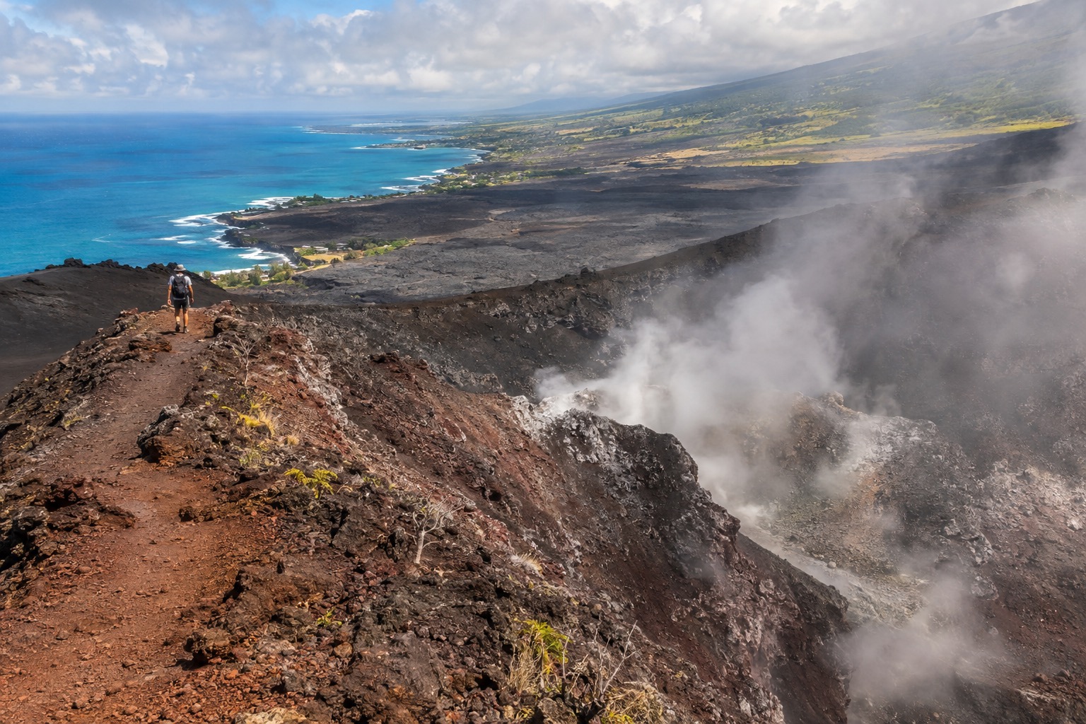 Steam rising from a volcanic crater rim trail under a partly cloudy sky