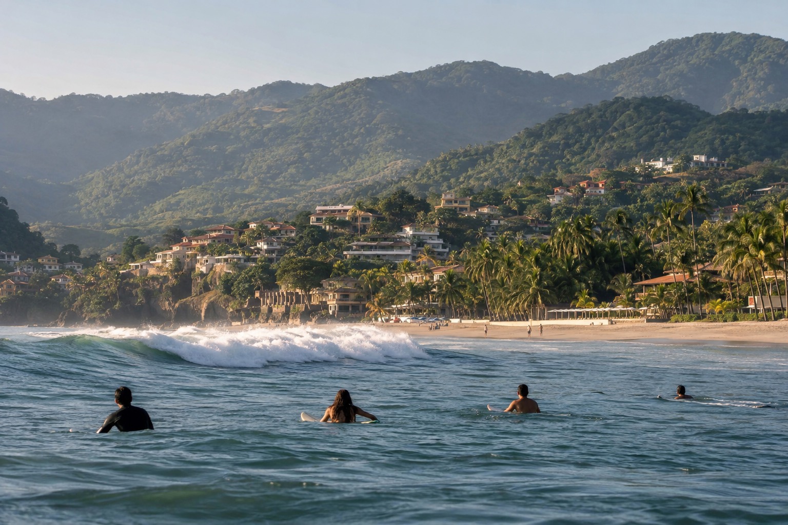 Surfers waiting for waves off a curved sandy town beach backed by palm trees on the Nayarit coast