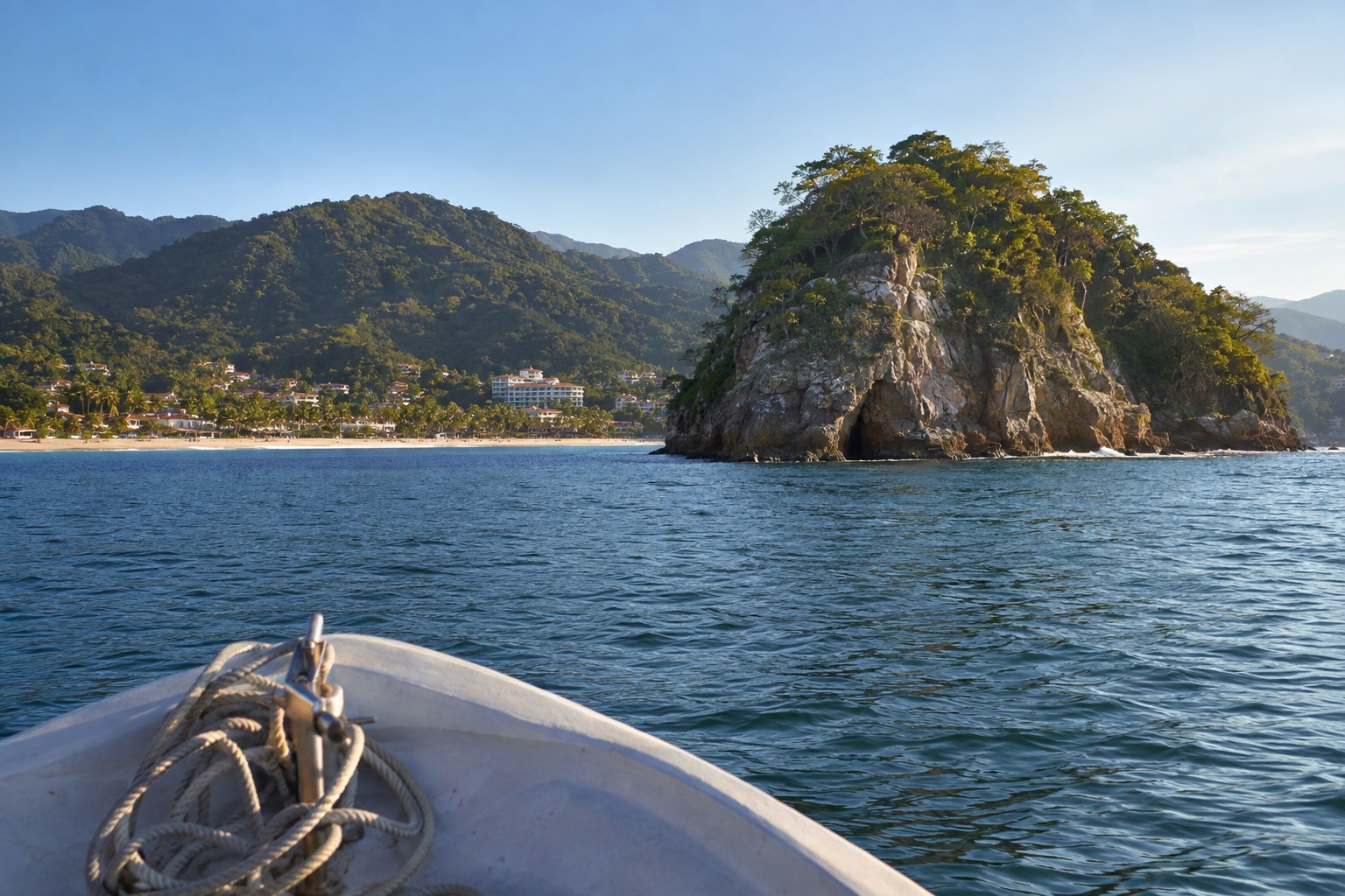 A small open boat crossing calm blue water toward a rocky island offshore under a clear sky