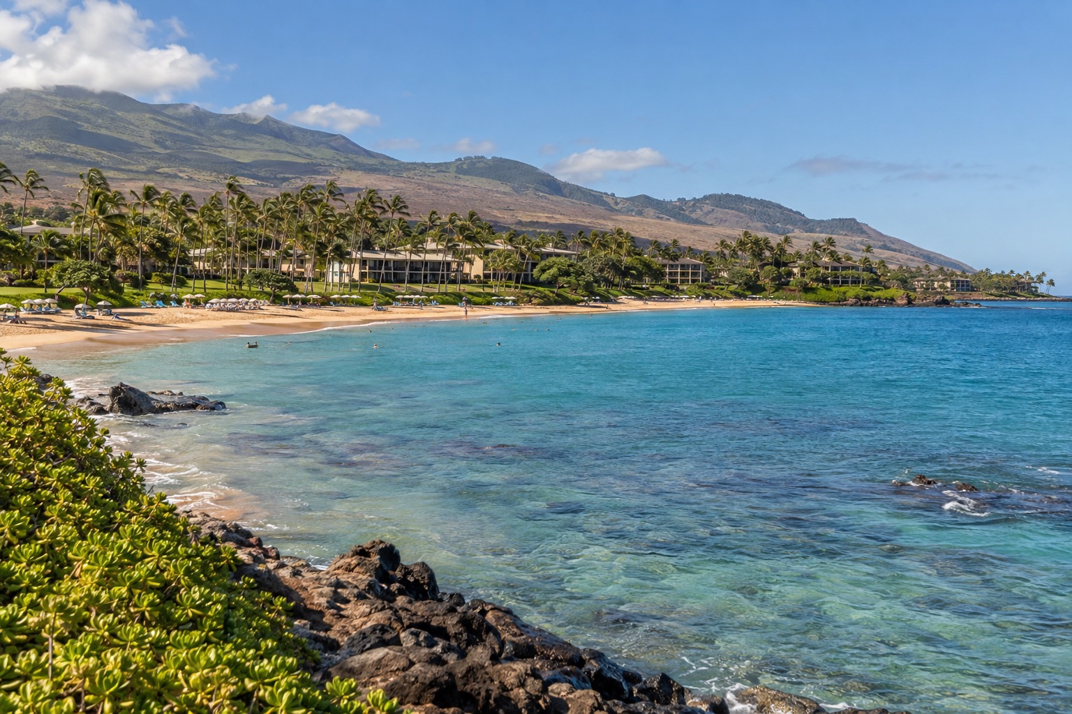 A crescent bay in Wailea with calm turquoise water fronting a row of low resort buildings among palm trees