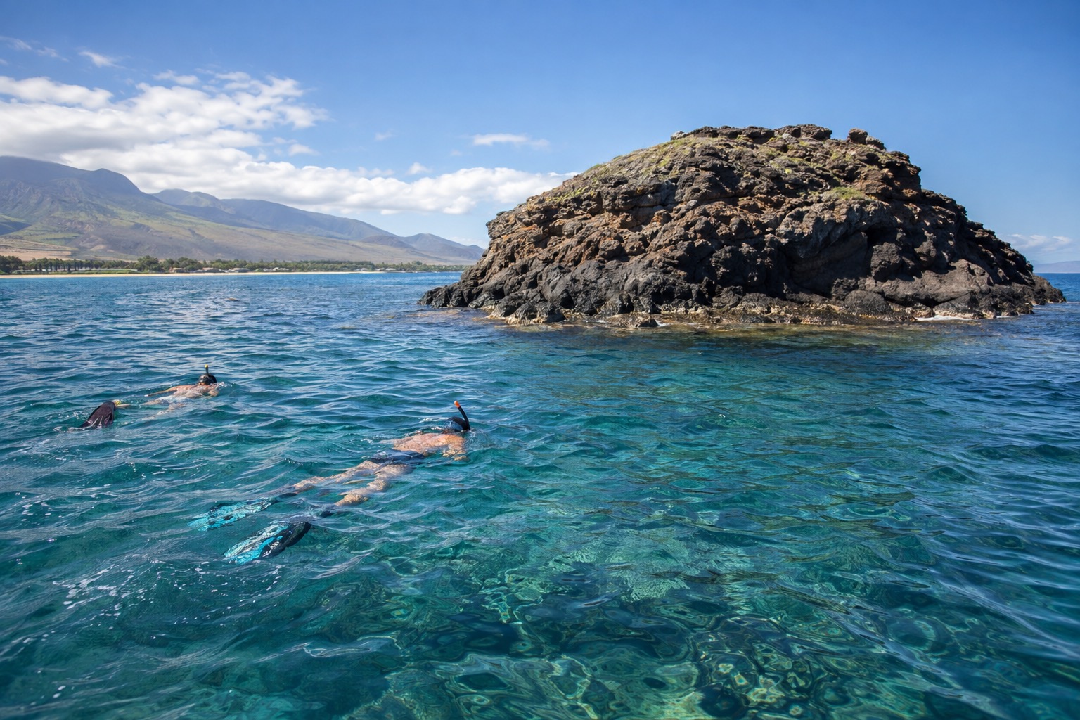 Snorkelers near a rocky crescent-shaped islet rising from blue water off Maui's coast