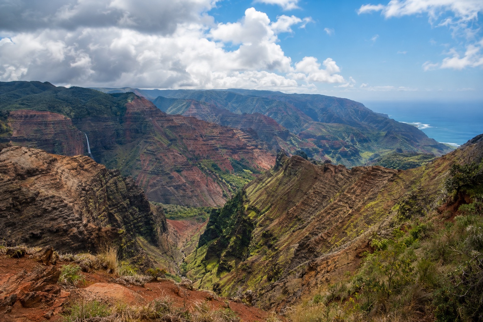 Layered red and green cliffs of Waimea Canyon seen from an overlook under a partly cloudy sky
